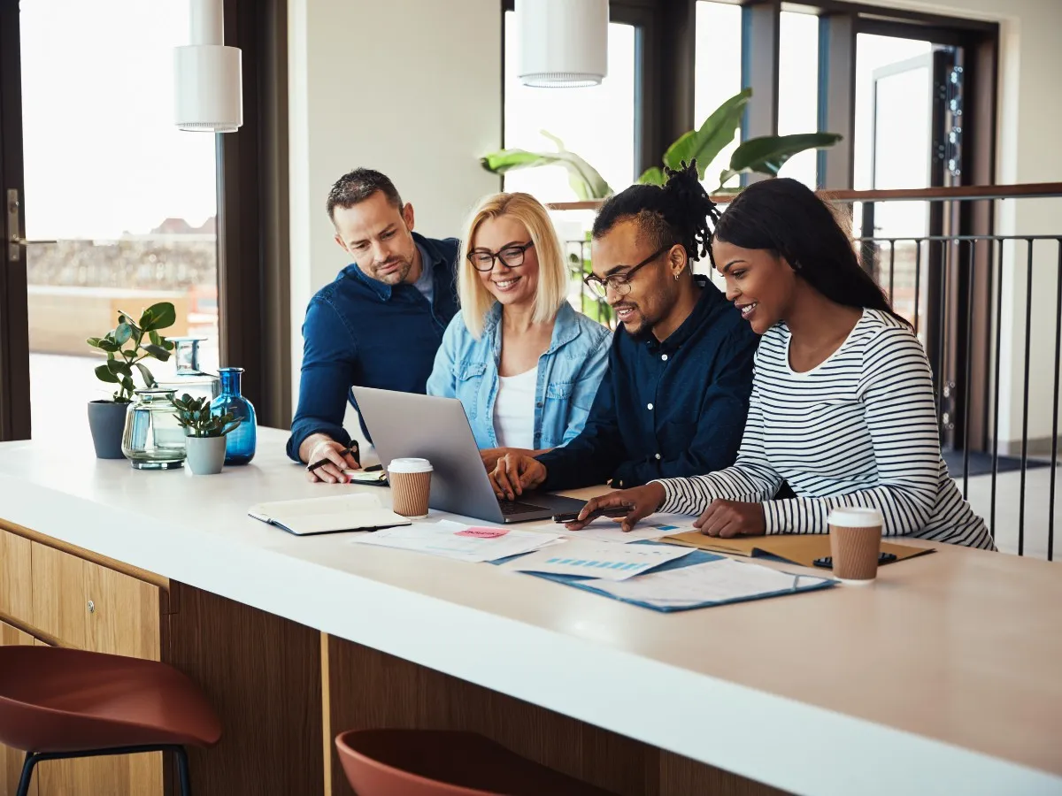 Four colleagues gathered around a laptop at a shared table, smiling and talking